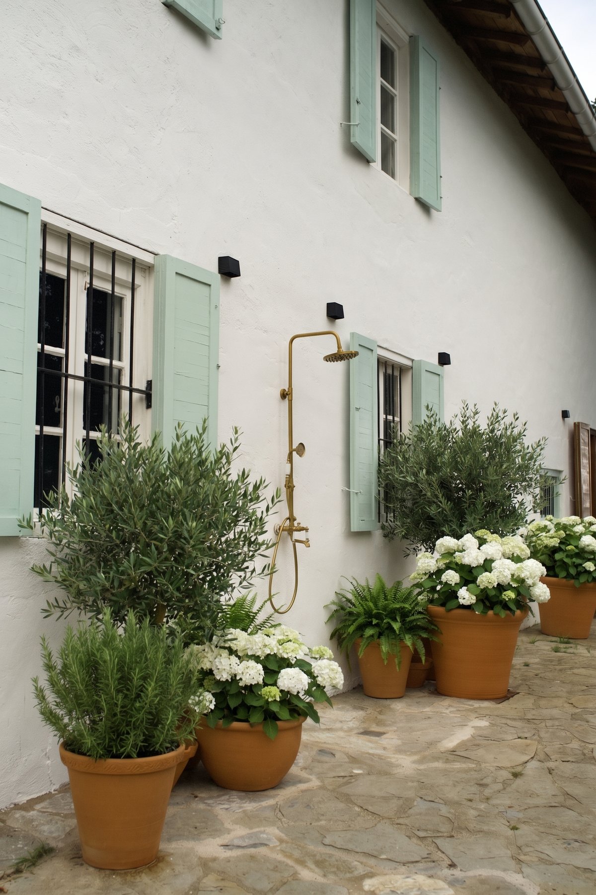 Ducha exterior de latón rodeada de plantas sobre la fachada encalada y contras verdes